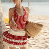 Woman in a red and white striped swimsuit holding a straw hat on a beach.